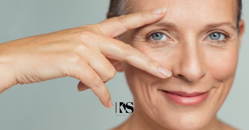 Silver haired woman highlights her eyes with fingers as she smiles at the camera. Overlay with black KDS Aesthetics logo.