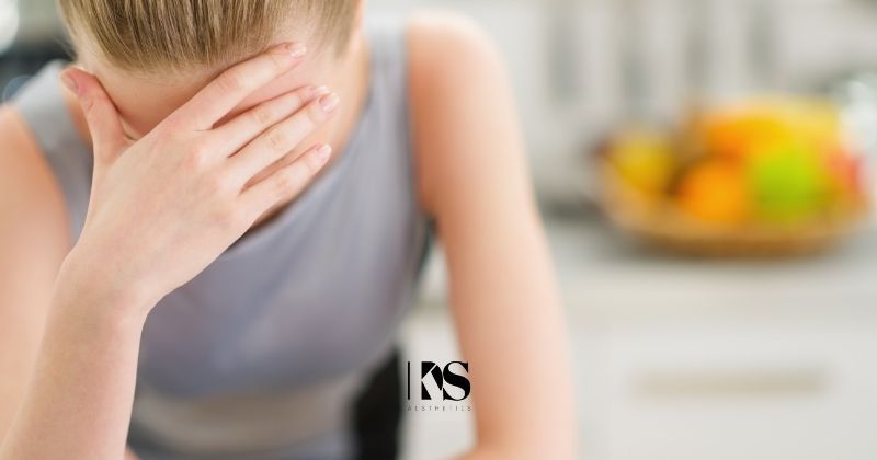 Blonde woman in gray dress looks down and holds head in pain from migraine headache contemplating if botox injections are a possible solution. KDS Aesthetics logo at bottom center in black. 